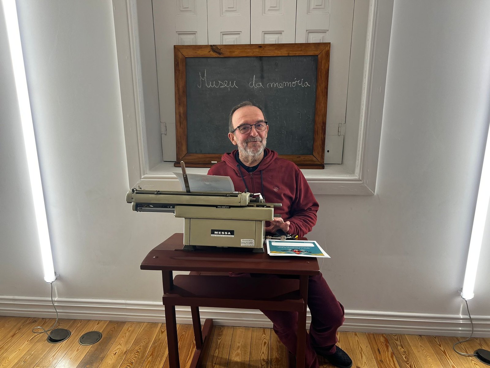 Homem sorrindo atrás de uma máquina de escrever antiga, com um quadro «Museu da memória» ao fundo